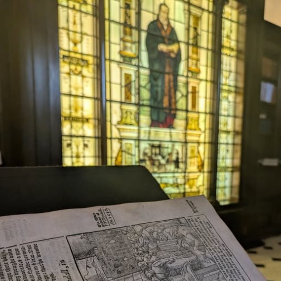 In foreground edge of early printed book, in background a stained glass window showing a man in 16th century clothes holding a book.