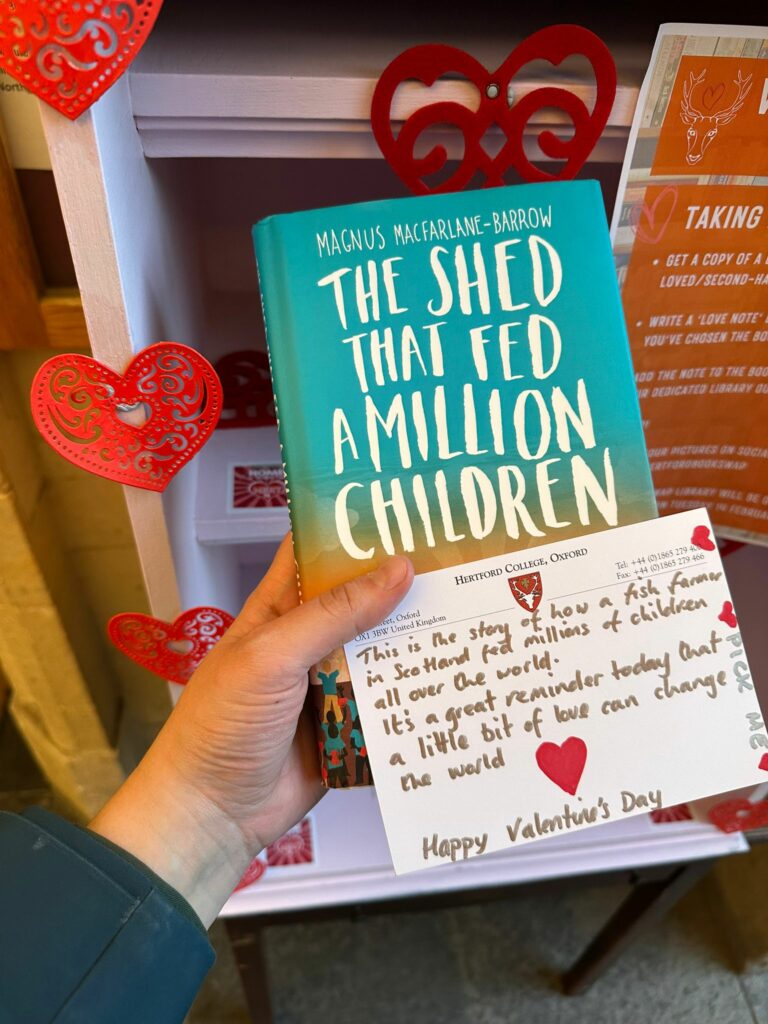 A student holds the book 'The shed that fed a million children' by Magnus Macfarlane-Barrow next to the Hertford Bookcase alongside a heartfelt dedication written on a card.