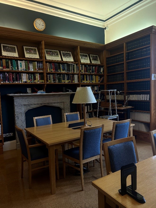 Reading room in Balliol College Library. Wooden study table with chairs, shelving in background with books and photographs on display. 