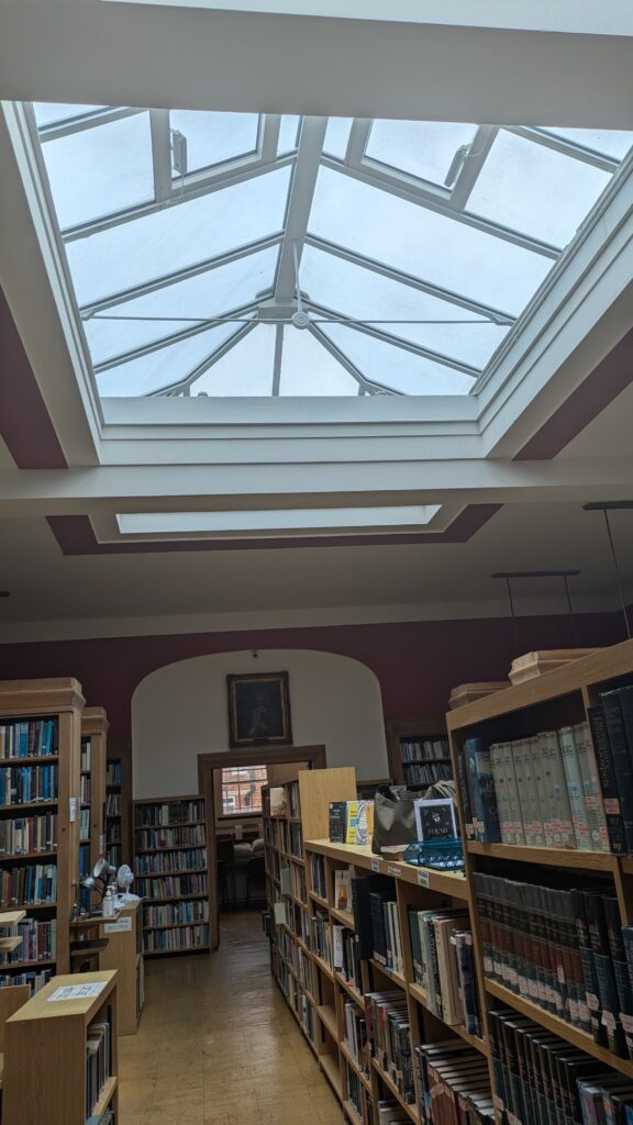 Inside of Regent's Park College Library, with low shelves down the middle of the room filled with books and a large skylight.