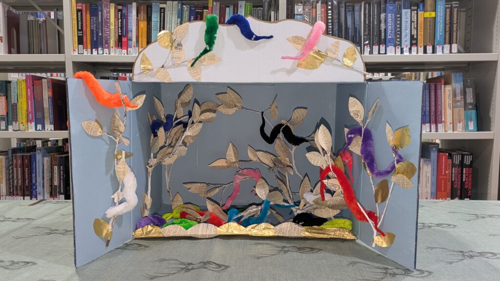 Worms wrapped around branches in an open box. Box is on a table with bookshelves in the background.