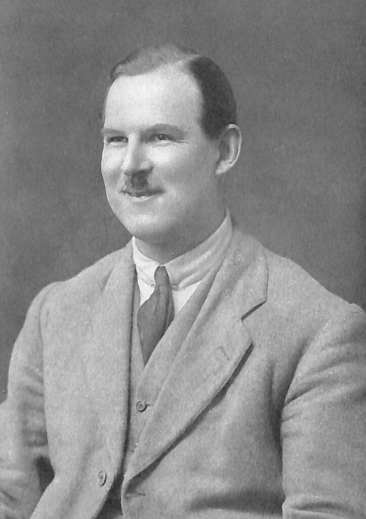 Black and white photograph of Principal Cruttwell. Image shows the head and upper body of a smiling white man in his 40s with neat short hair and moustache, wearing light coloured a suit.