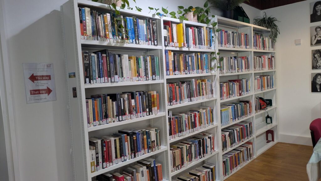 A row of book cases filled with library books. The books are very neatly arranged. Around the edges of the shelves are houseplants, some portraits and directional signage.