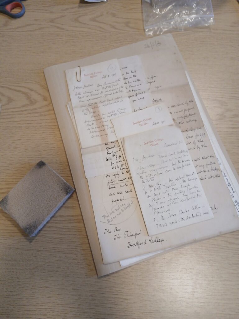 A bundle of archival handwritten letters on a wooden desk, with a cleaning sponge next to them.